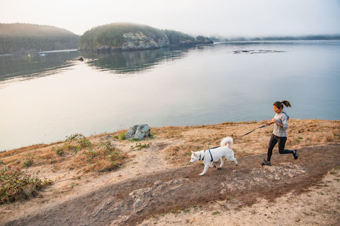 Woman running with a dog on a trail by a lake with mountains in the background