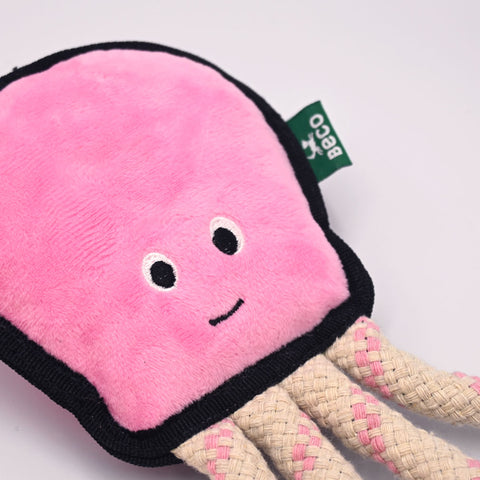 Pink plush toy with a smiley face and rope, featuring a visible brand tag on a white background. Detail view.