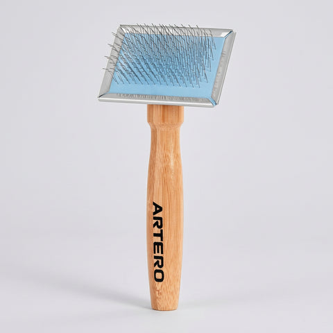 An ARTERO pet grooming brush with a light-colored wooden handle and a metal head featuring a light blue protective cover and silver pins, shown on a solid white background.