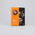 A boxed pet safety light product shown on a black background with a floating box and soft shadow. The package is orange and black, with a circular cutout showing the red safety light and a black dog wearing the light.