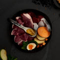 Assorted raw meats, vegetables, and a boiled egg on a dark surface with a spoon.