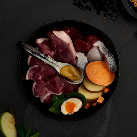 Assorted raw meats, vegetables, and a boiled egg on a dark surface with a spoon.