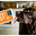 A chocolate brown dog licking a white carton of Kaniules Bone Broth, with the carton's orange and blue label clearly visible against a blurred indoor background.