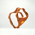 An orange dog harness with patterned straps and a yellow logo patch, shown floating against a white background.
