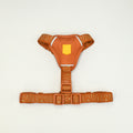 An orange dog harness with a matching adjustable waist belt, featuring a subtle tonal pattern and a small yellow shield logo on the chest plate, set against a plain off-white background.