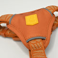 Close-up of an orange pet harness with a woven orange-and-beige strap, gray trim, and a yellow square brand patch on the center.